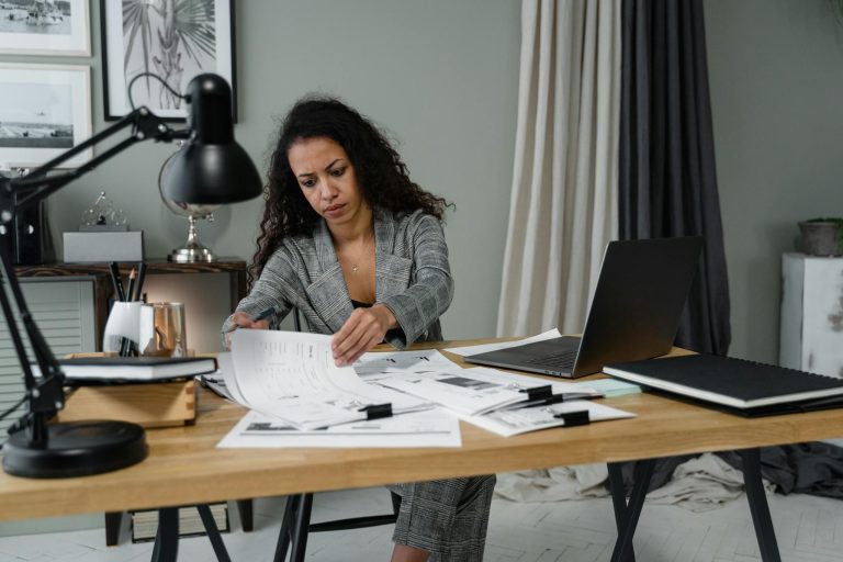 Businesswoman in office reviewing documents at desk with laptop and lamp.